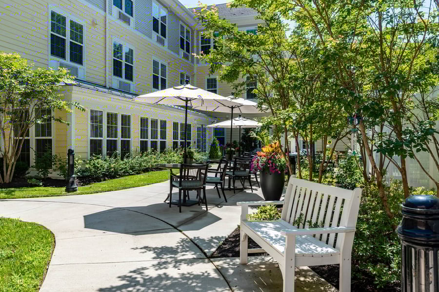 A serene outdoor courtyard with shaded patio seating, lush trees, and colorful flowers surrounding the grounds of our senior living community in Gambrills, MD.