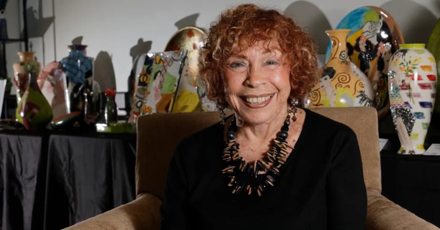 Smiling older woman with curly red hair wearing a black top and a bold gold statement necklace, seated indoors with colorful artwork in the background.
