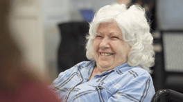 A resident with soft white curls smiles brightly during a group gathering, her joy lighting up the room at Arbor Terrace Prince William Commons, a warm senior living community in Woodbridge VA.