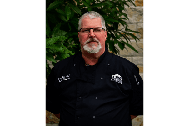 A culinary staff member in a black chef's coat and Arbor Company logo poses in front of greenery — introducing the dining team at senior living Lakeway Texas.