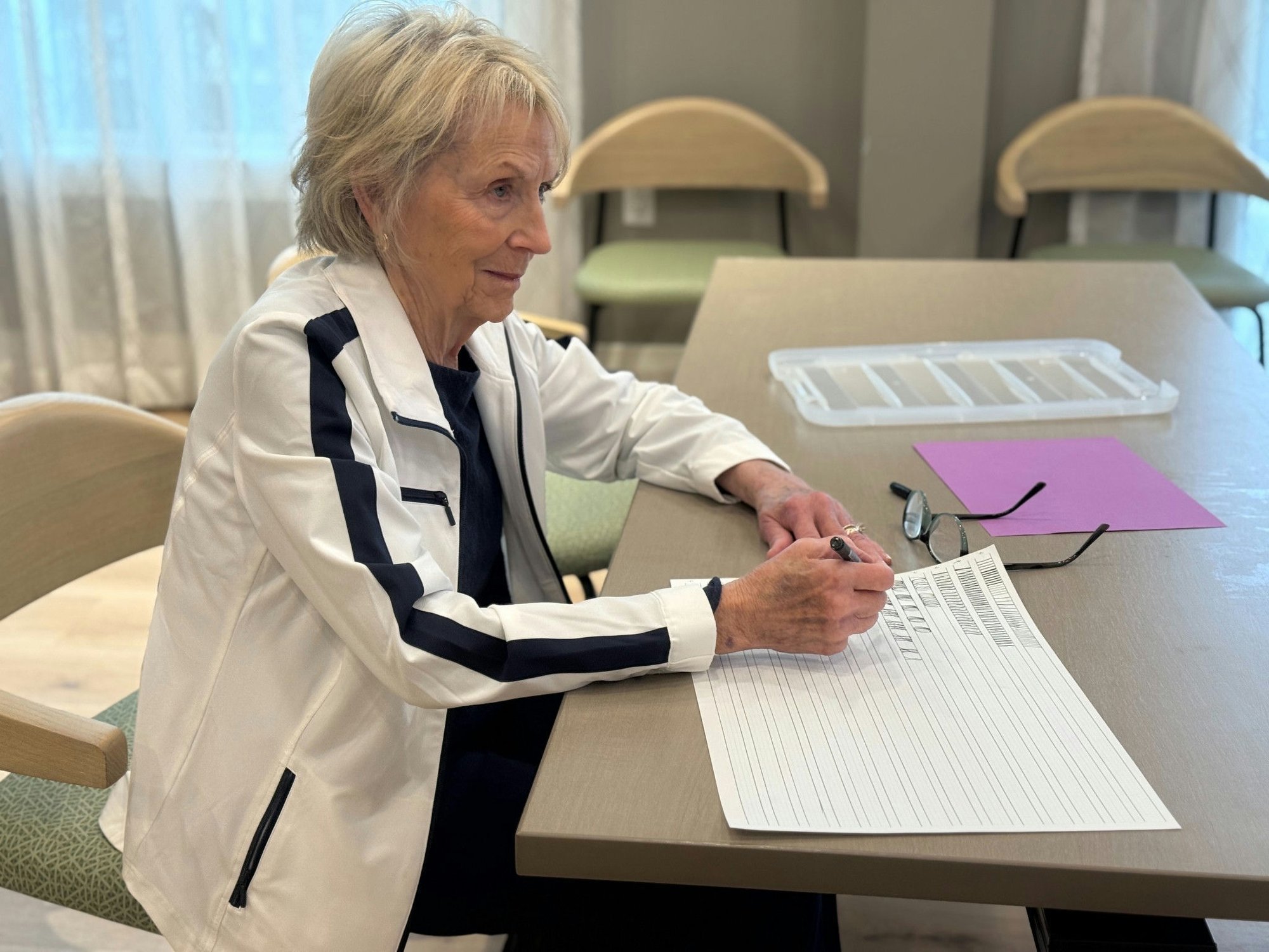 A resident seated at a table at Opus East Memphis in East Memphis, TN, focused on writing with a pen on lined paper, with a pink folder and glasses resting nearby, enjoying a quiet activity at this senior living community.