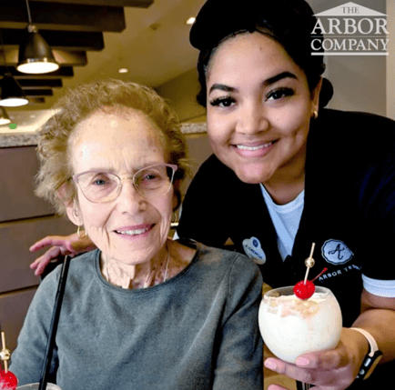 An Arbor Terrace staff member beams alongside a resident who smiles warmly for the camera, with an ice cream sundae in the foreground, showcasing the joyful, people-first spirit of assisted living Woodbridge VA.