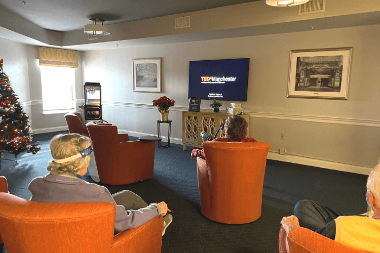Residents relax in cozy armchairs watching a TEDx talk in a warmly decorated lounge with a popcorn machine and holiday tree, showcasing the engaging senior living with dining and activities Herndon VA offers at Arbor Terrace Herndon.