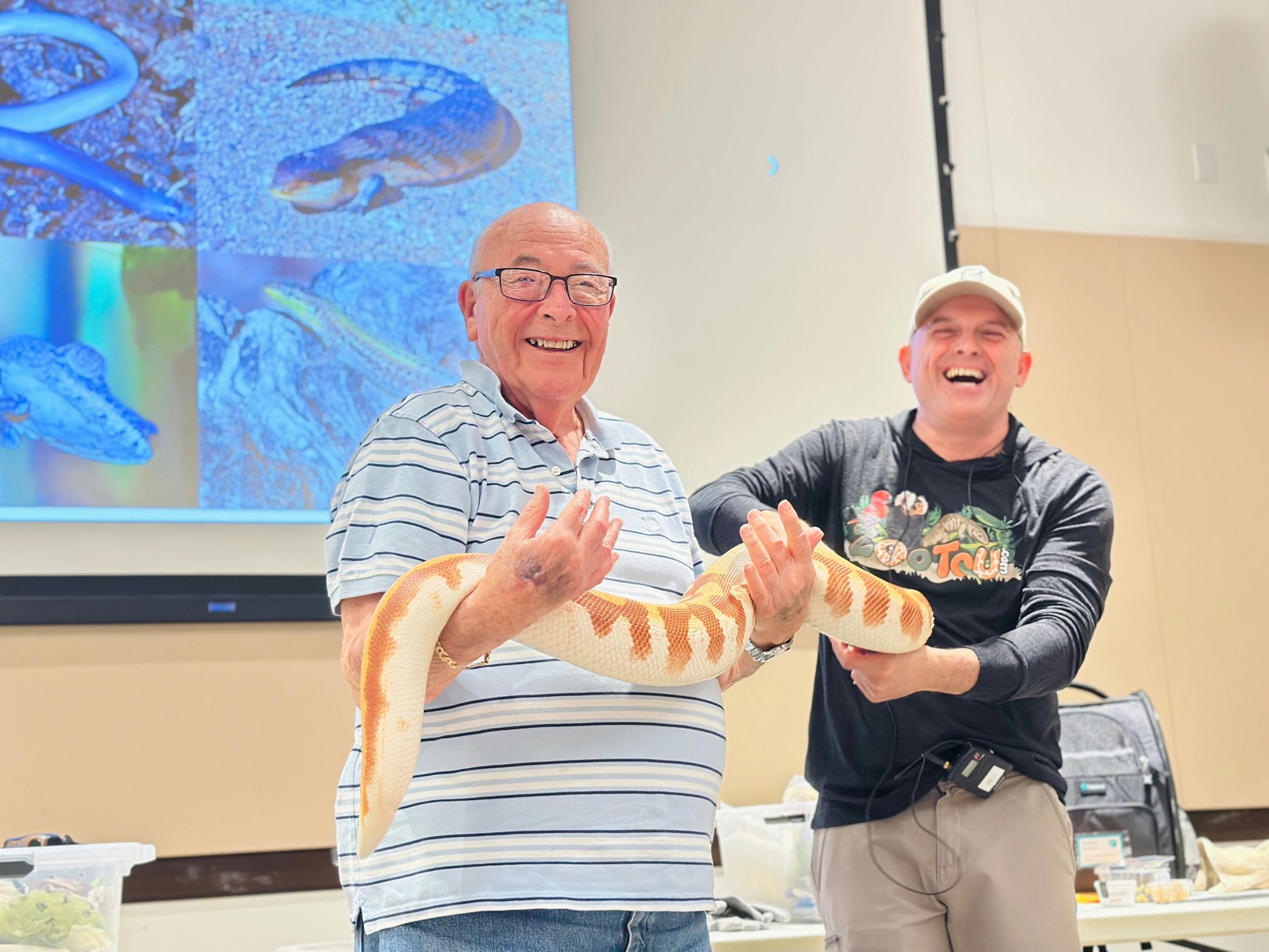 Two men laugh while holding a snake presented during a wildlife program, enjoying a fun and engaging activity as part of an active senior lifestyle at The Arbor Company.