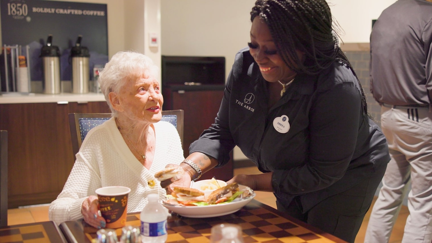 A dining staff member smiles warmly while serving a fresh meal to a resident at Arbor Terrace Waugh Chapel, reflecting the attentive, resort-style dining experience and personalized care that define senior living at this assisted living community in Gambrills, MD.